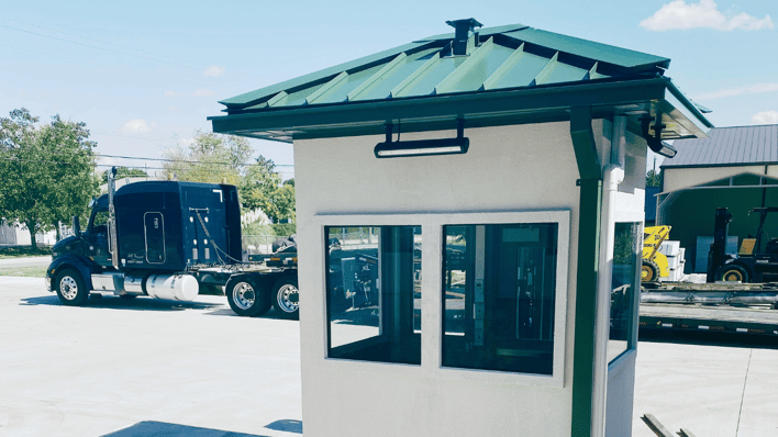 A modern guard booth with a green metal roof, large windows, and a secure design, positioned near a parked semi-truck in an industrial setting.