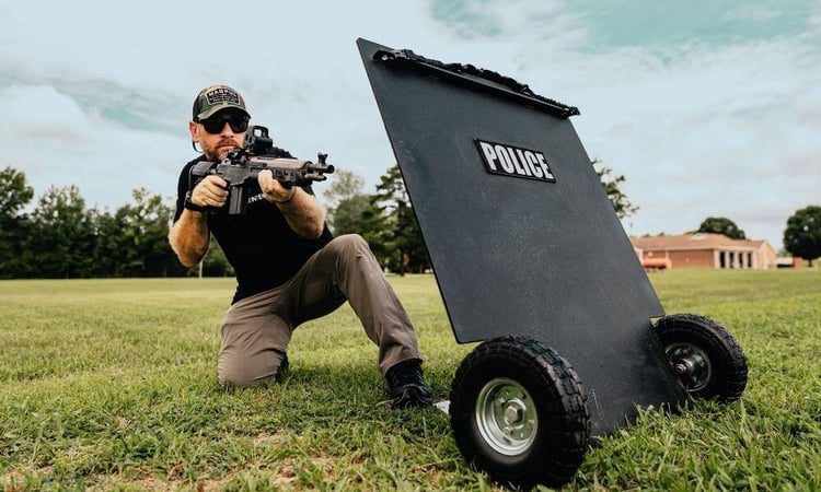 A tactical operator kneeling with a rifle beside a mobile ballistic shield equipped with wheels, used for law enforcement defense and field operations.